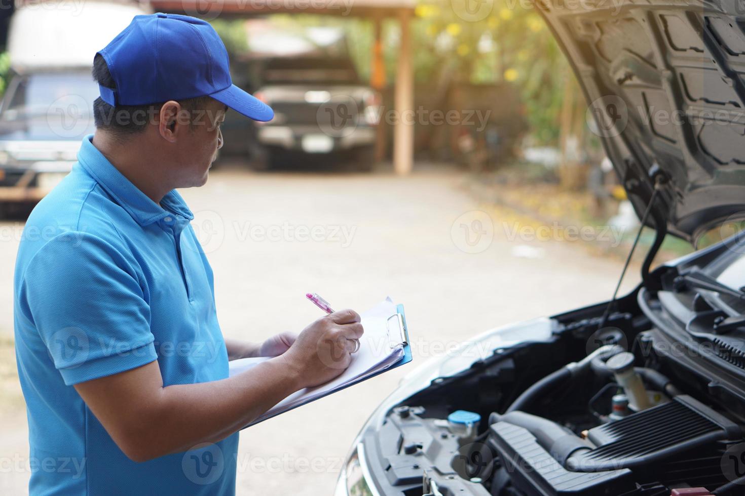 Aziatisch Mens monteur draagt blauw pet en blauw shirt, houdt papier, controle en analyseren auto motor onder de kap. concept, buitenshuis auto inspectie onderhoud. beweren voor ongeluk verzekering. foto