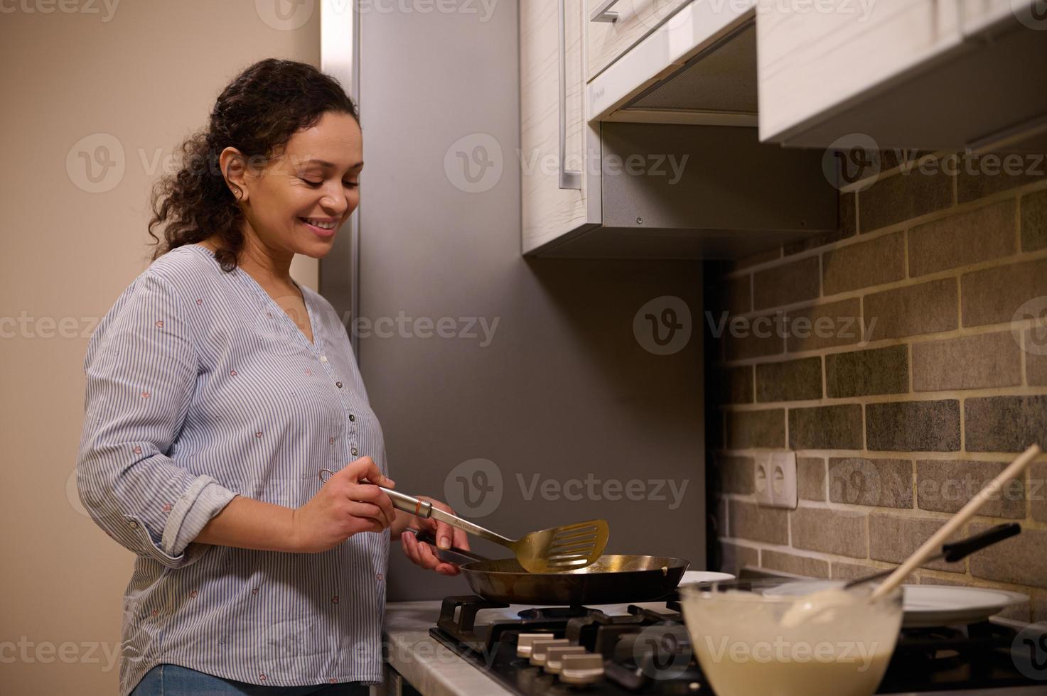 Afrikaanse Amerikaans vrouw, gelukkig huisvrouw kookt pannekoeken in een frituren pan, glimlacht zoet, staand in de buurt de zwart fornuis in de huis keuken foto