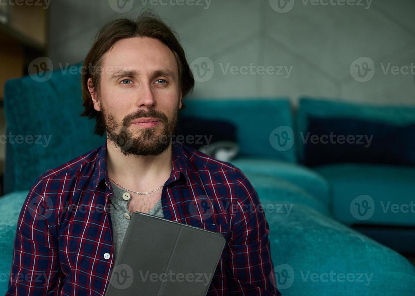 portret van een knap Kaukasisch Mens Holding een digitaal tablet in zijn handen en vol vertrouwen op zoek Bij camera, zittend Aan een blauw sofa Bij huis foto