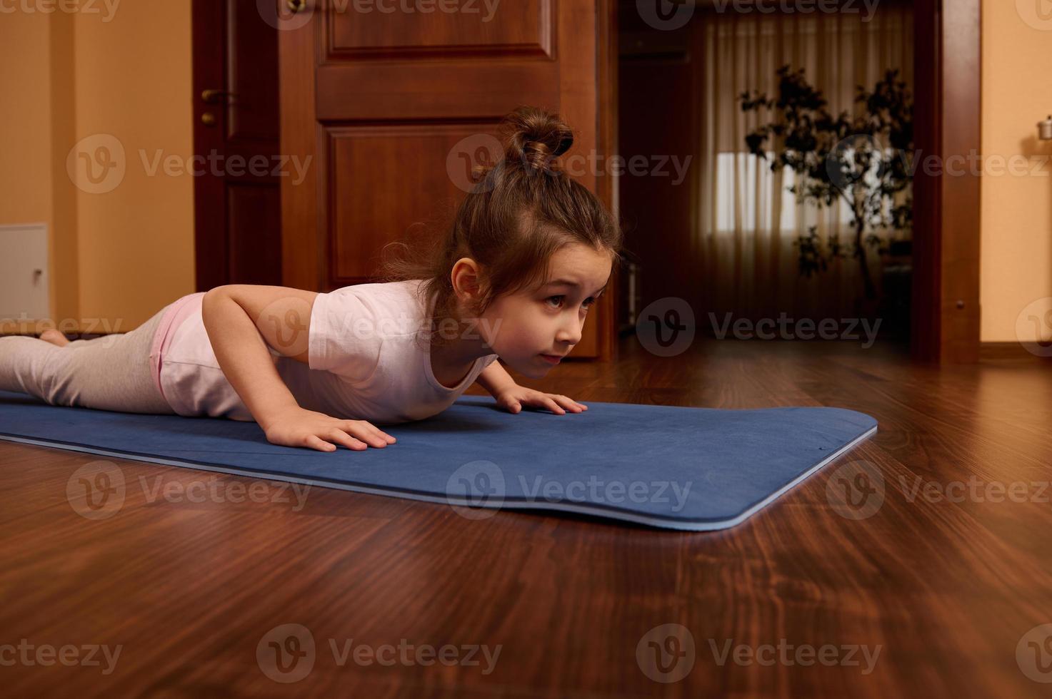 vastbesloten weinig meisje aan het doen Duwen opstaan, vier ledematen personeel oefening, blijven in plank Aan mat terwijl sporten binnenshuis foto