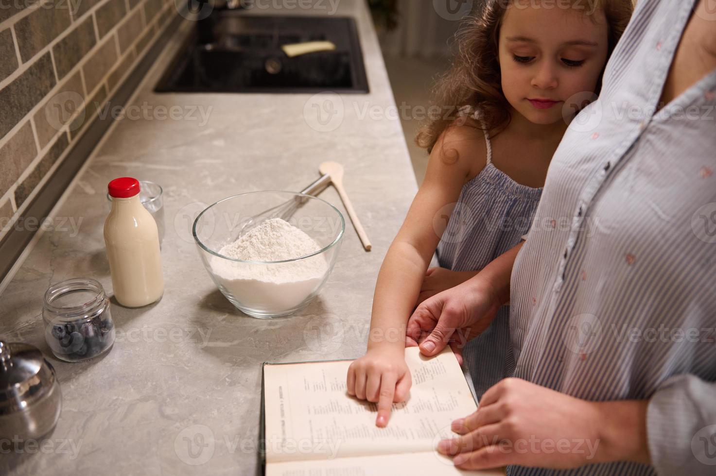 mooi gelukkig kind, aanbiddelijk weinig meisje points met haar vinger Aan een recept boek, staand Bij keuken aanrecht dichtbij naar haar mam terwijl genieten van Koken samen, voorbereidingen treffen deeg voor vasten dinsdag foto