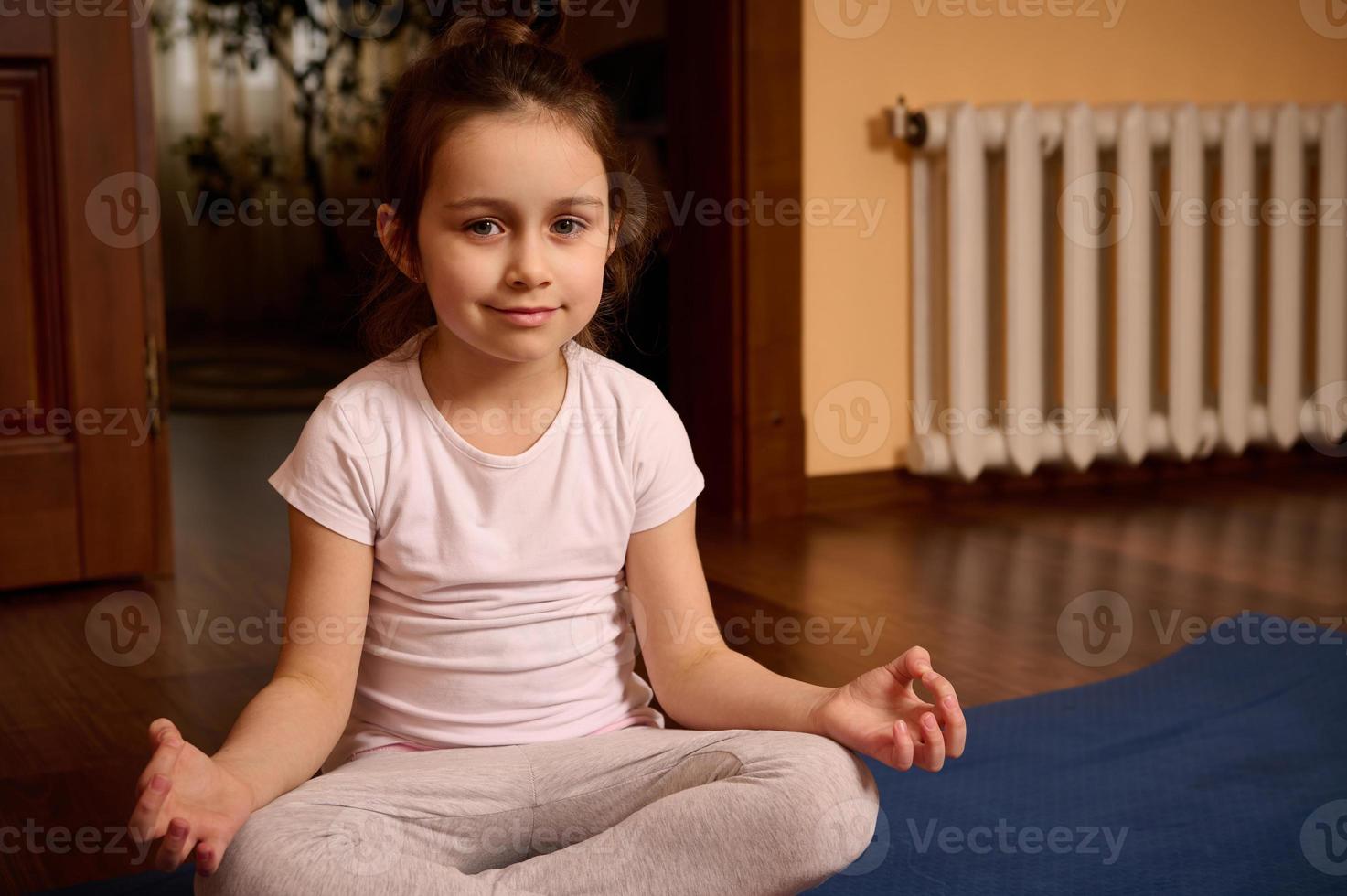 mooi Europese weinig meisje in sport- uniform, glimlachen op zoek Bij camera terwijl zittend in lotus positie Aan yoga mat. foto