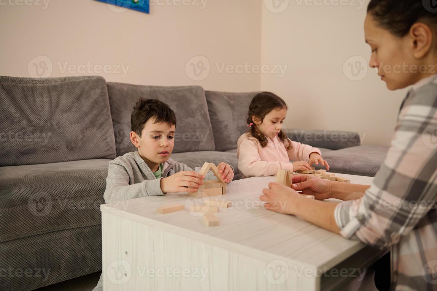 focus Aan een knap school- leeftijd jongen gebouw houten huis met blokken en bakstenen, zittend Bij tafel in de buurt haar zus en mam gefocust Aan bouw van structuren. familie tijdverdrijf en vrije tijd concept foto