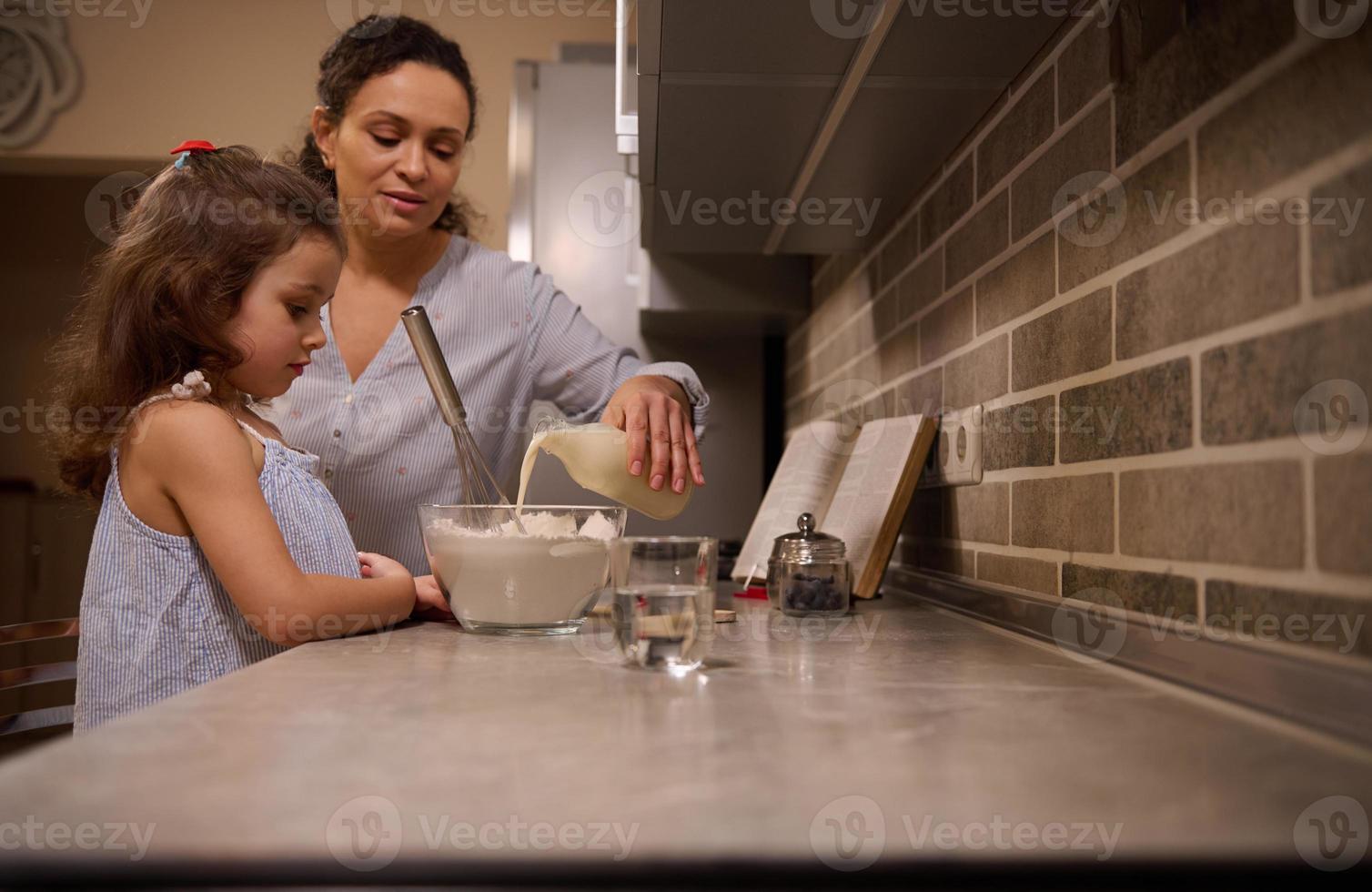 levensstijl portret van een mam gieten melk in kom van meel en menging ingrediënten met een vliegenmepper naar bereiden pannenkoek deeg. moeder en dochter - mooi kind zijn Koken samen in de huis keuken foto