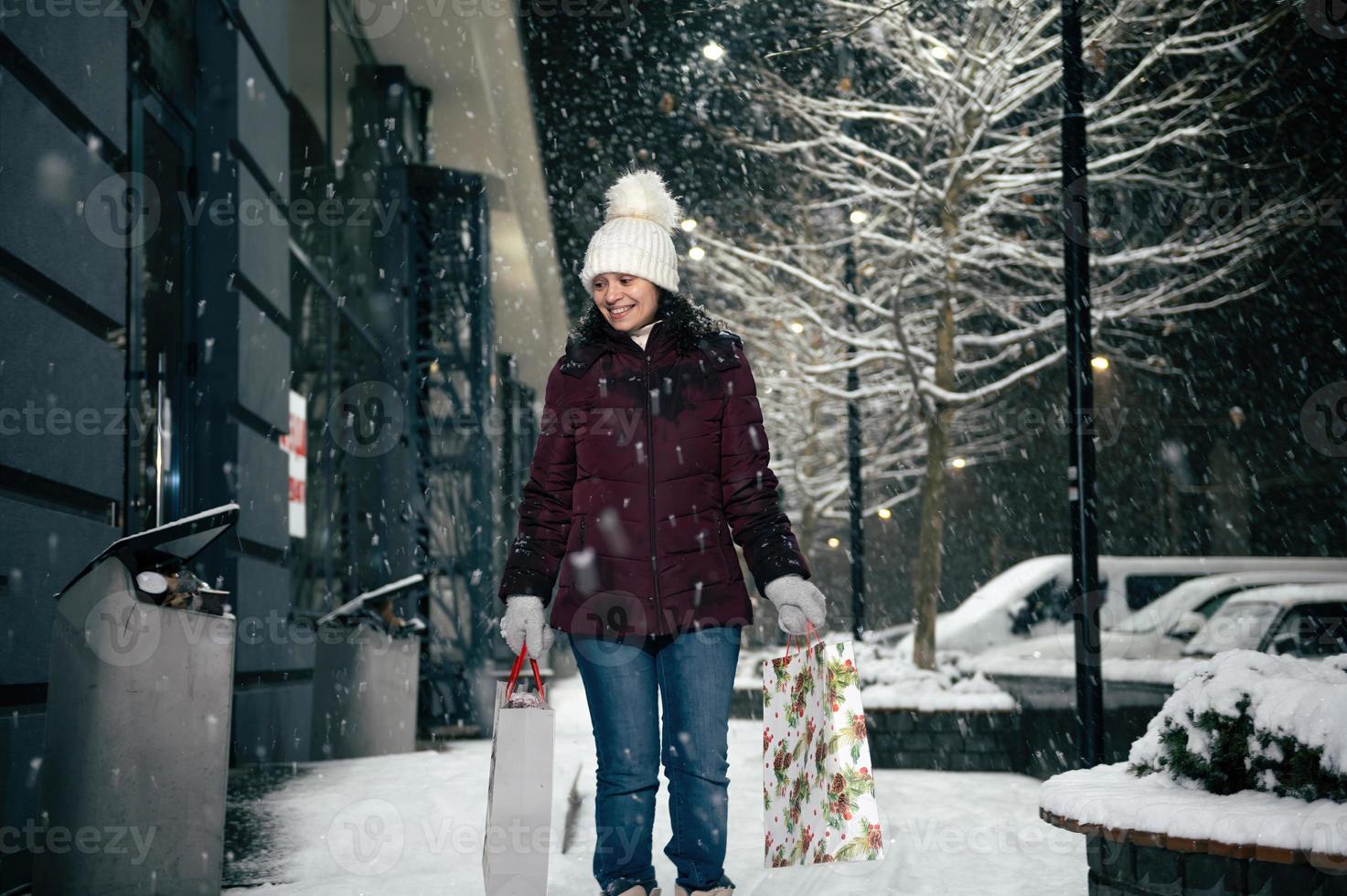 verrukkelijk jong vrouw wandelen naar beneden de straat met boodschappen doen Tassen met Kerstmis geschenk doos in haar handen. vrolijk kerstmis. foto