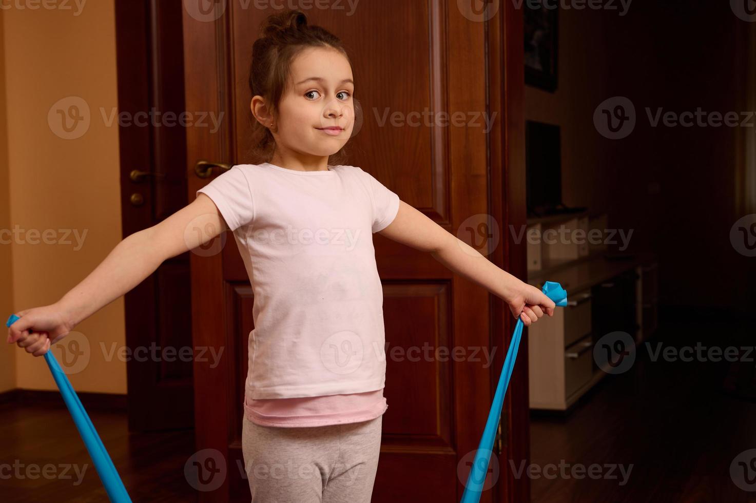 Kaukasisch schattig weinig meisje gebruik makend van elastisch rubber band, aan het doen lichaamsgewicht , uitrekken opdrachten, glimlachen op zoek Bij camera foto