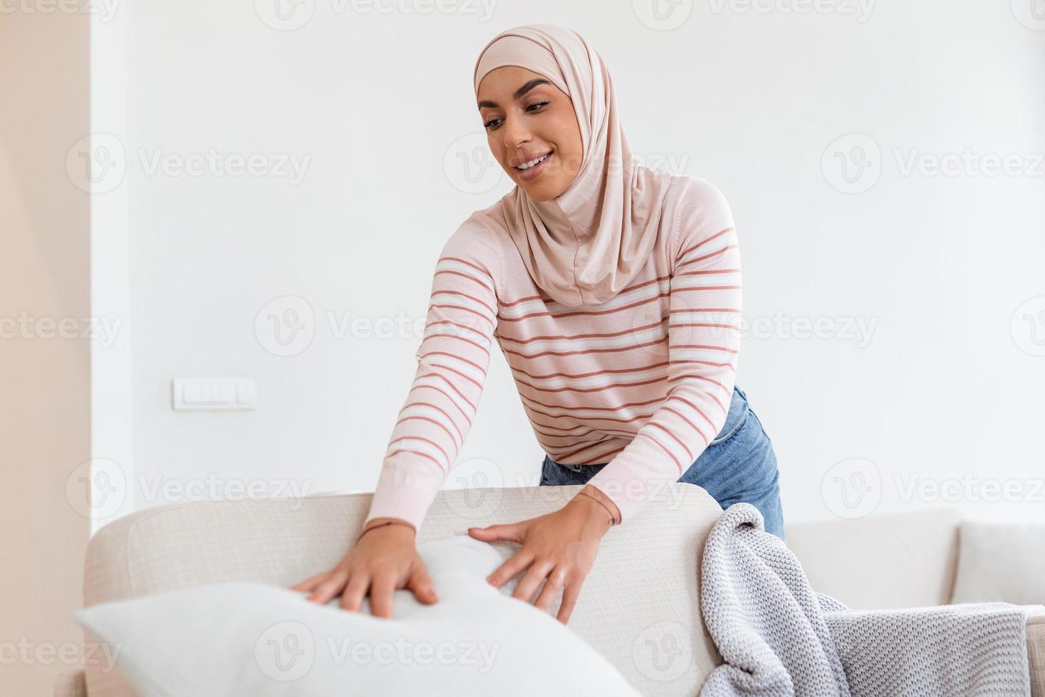 lief jong Arabisch moslim vrouw zetten zacht kussens en plaid Aan comfortabel bank, maken haar huis knus en warm foto
