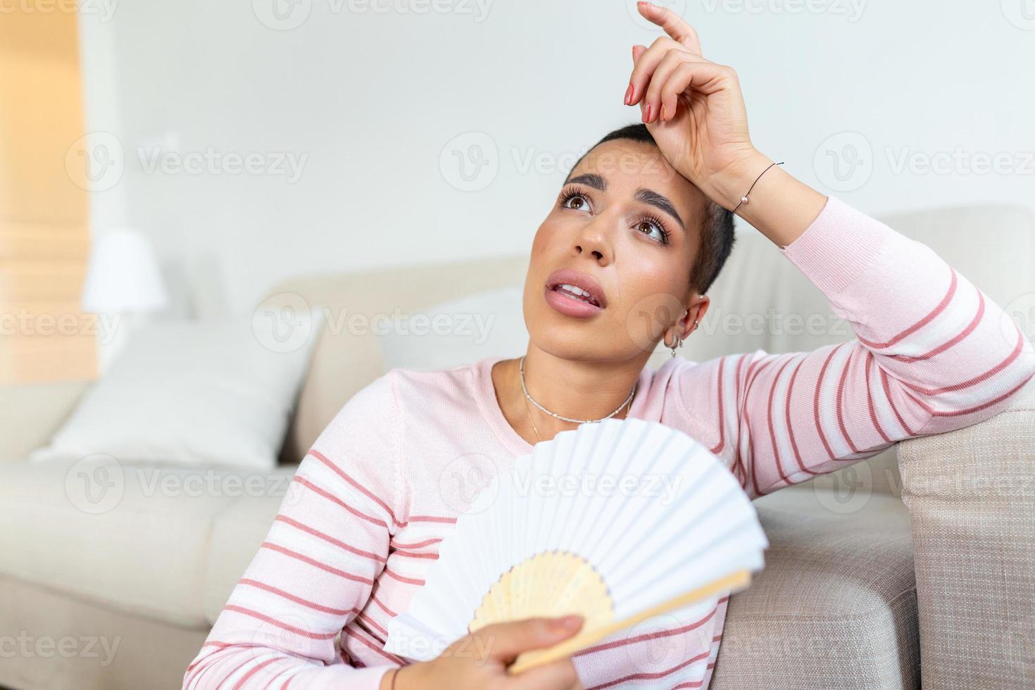 oververhit vrouw zitten Aan bankstel Bij huis voelen warm golvend met hand- ventilator koeling omlaag, zweten meisje kom tot rust Aan sofa in leven kamer houden wankelen lijden van warmte, Nee lucht conditioner systeem foto