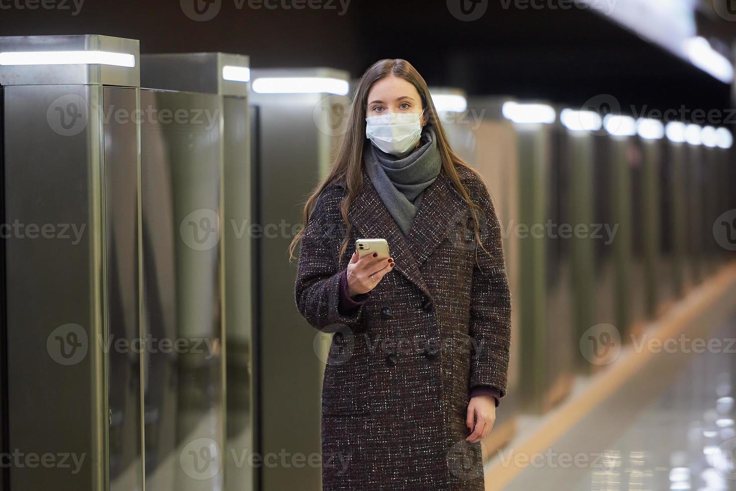 een vrouw met een medisch gezichtsmasker wacht op een trein en houdt een smartphone vast foto