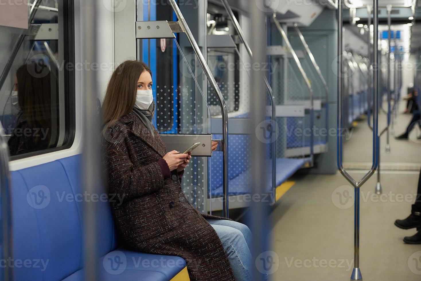 een vrouw met een gezichtsmasker zit en gebruikt een smartphone in een moderne metro foto