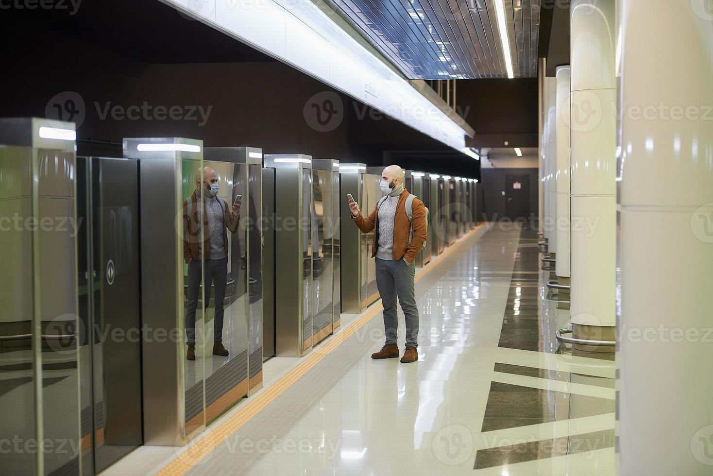 een man met een gezichtsmasker gebruikt een smartphone terwijl hij op een metro wacht foto