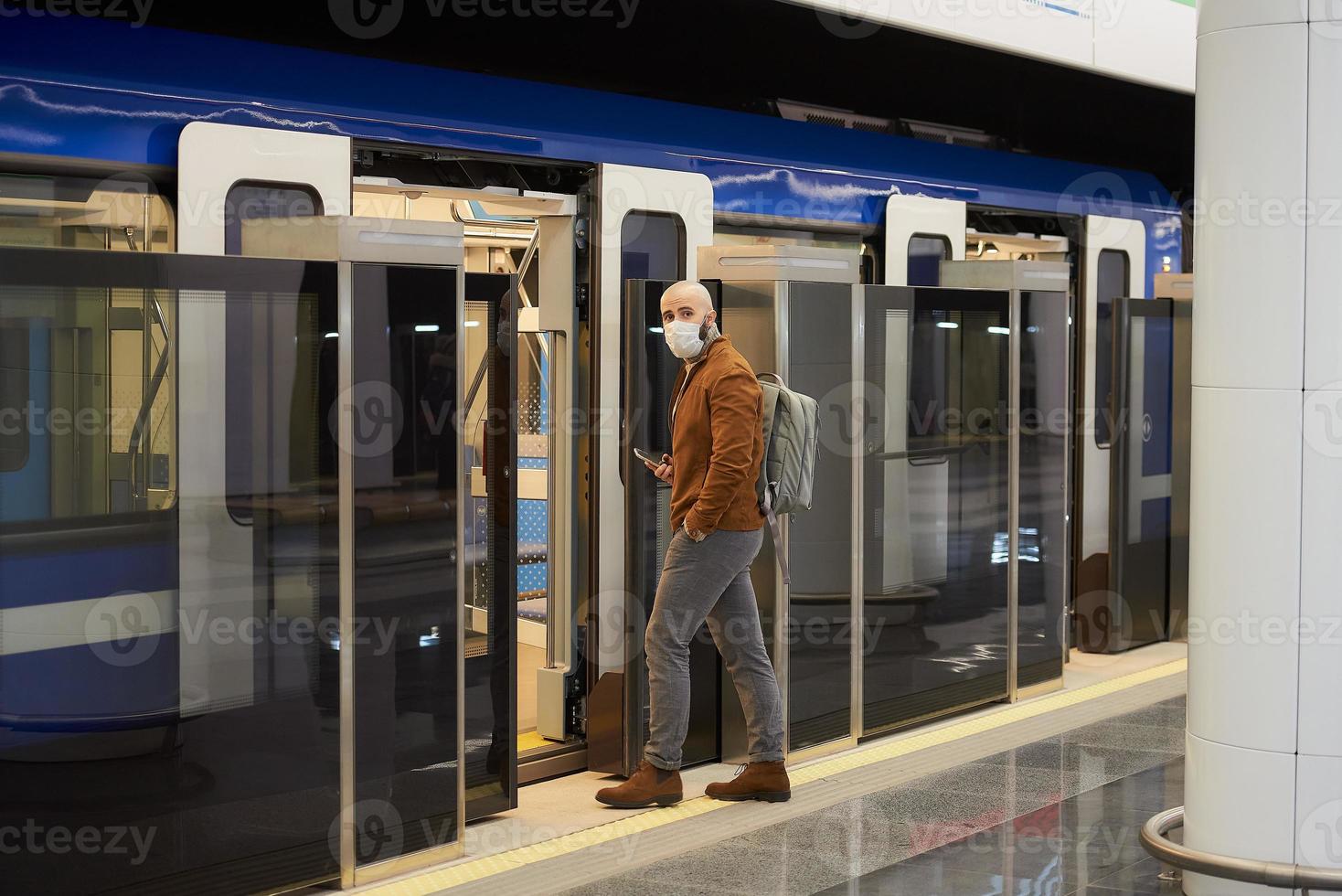 een man met een medisch gezichtsmasker houdt een smartphone vast bij het betreden van een metro foto