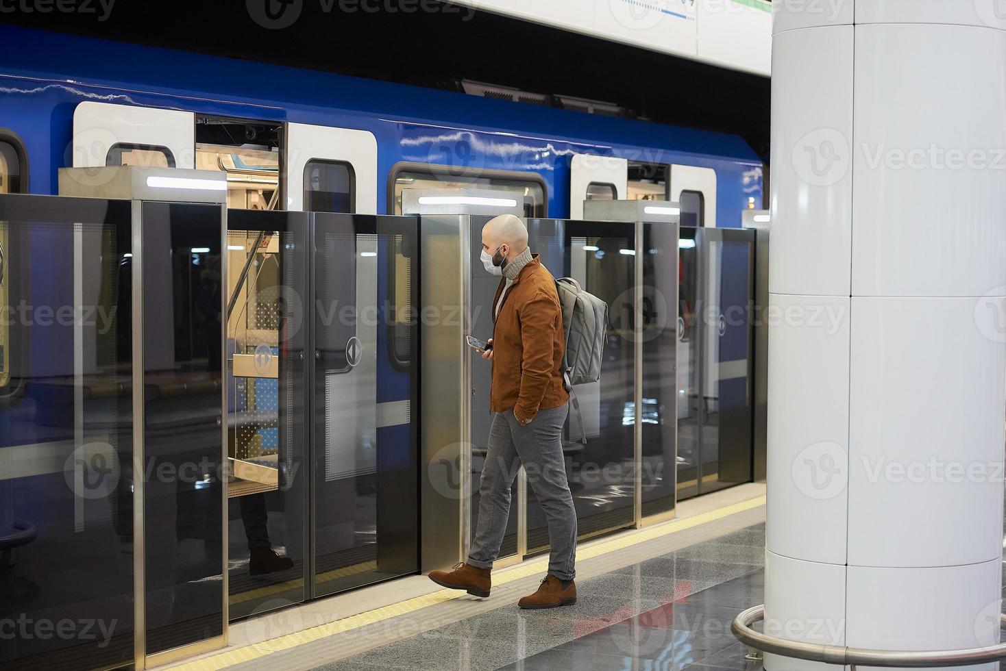 een man met een medisch gezichtsmasker houdt een smartphone vast bij het betreden van een metro foto