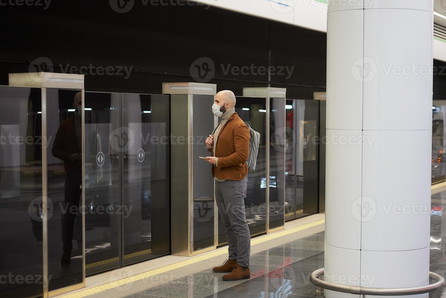 een man met een gezichtsmasker gebruikt een smartphone terwijl hij op een metro wacht foto