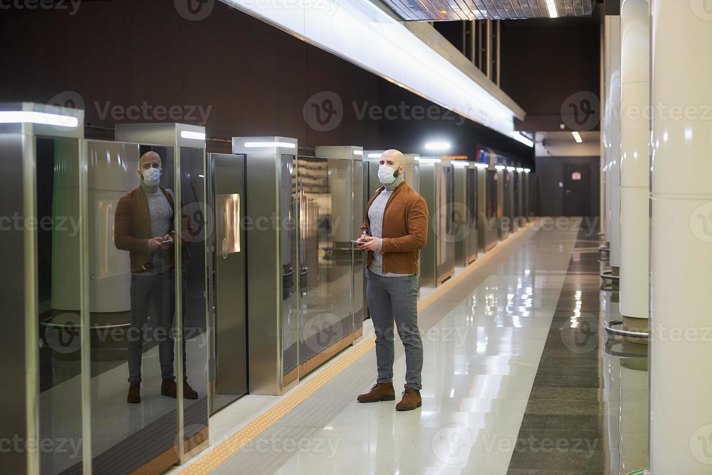 een man met een gezichtsmasker houdt een smartphone vast terwijl hij op een metro wacht foto