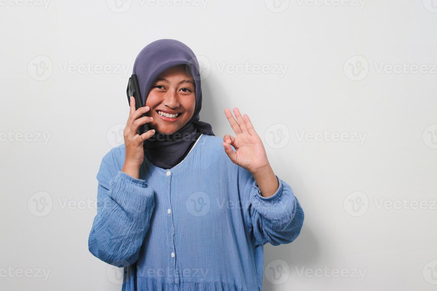 gelukkig jong Aziatisch meisje in hijab gekleed in gewoontjes overhemd zeggen Hallo naar de camera terwijl pratend Aan telefoon foto