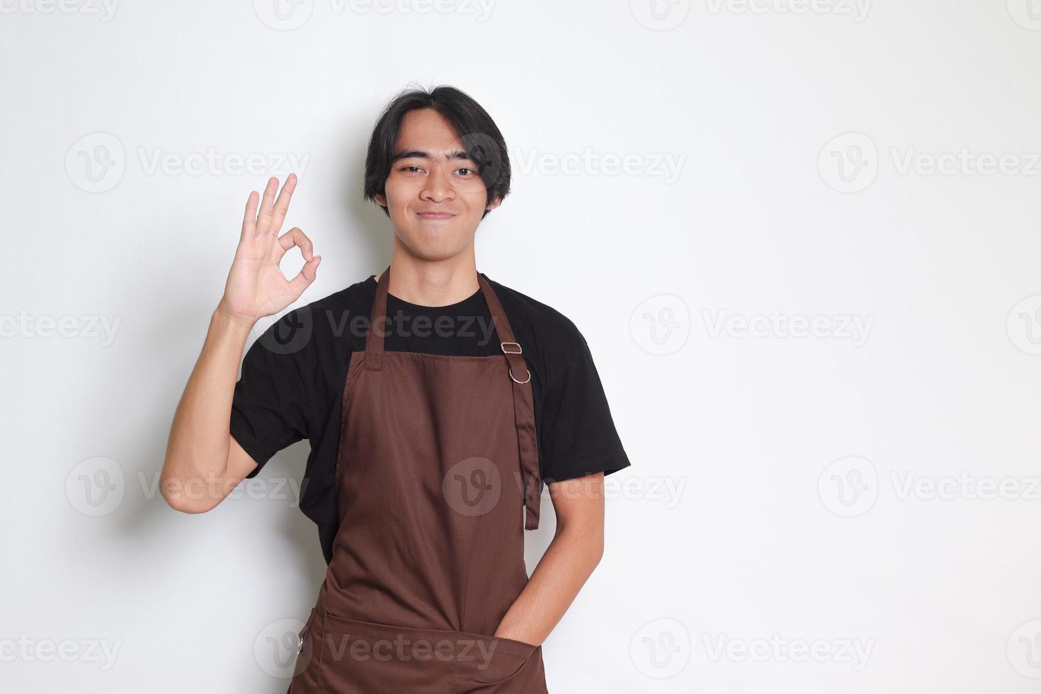 portret van aantrekkelijk Aziatisch barista Mens in bruin schort tonen OK hand- gebaar en glimlachen op zoek Bij camera. reclame concept. geïsoleerd beeld Aan wit achtergrond foto