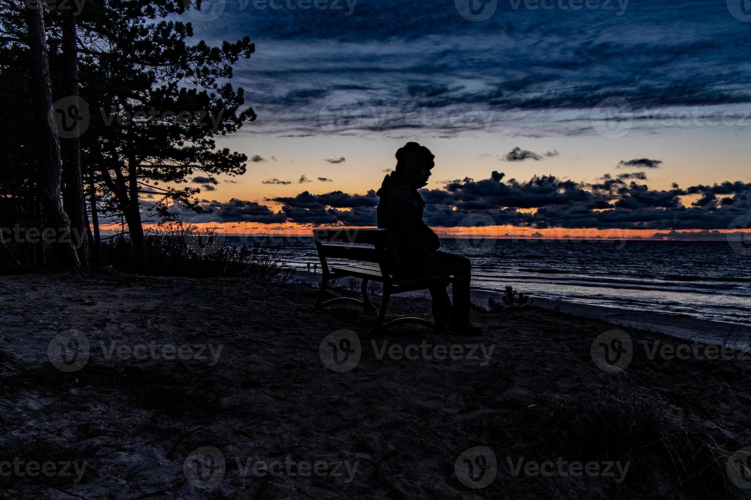 kleurrijk zonsondergang Aan de zee opgemerkt door een Mens in een pet zittend Aan een zwart bank Aan een duin foto