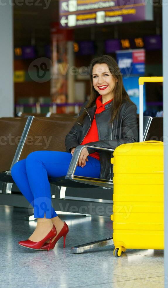 vrouw in helder kleren met geel koffer zit Bij de luchthaven foto