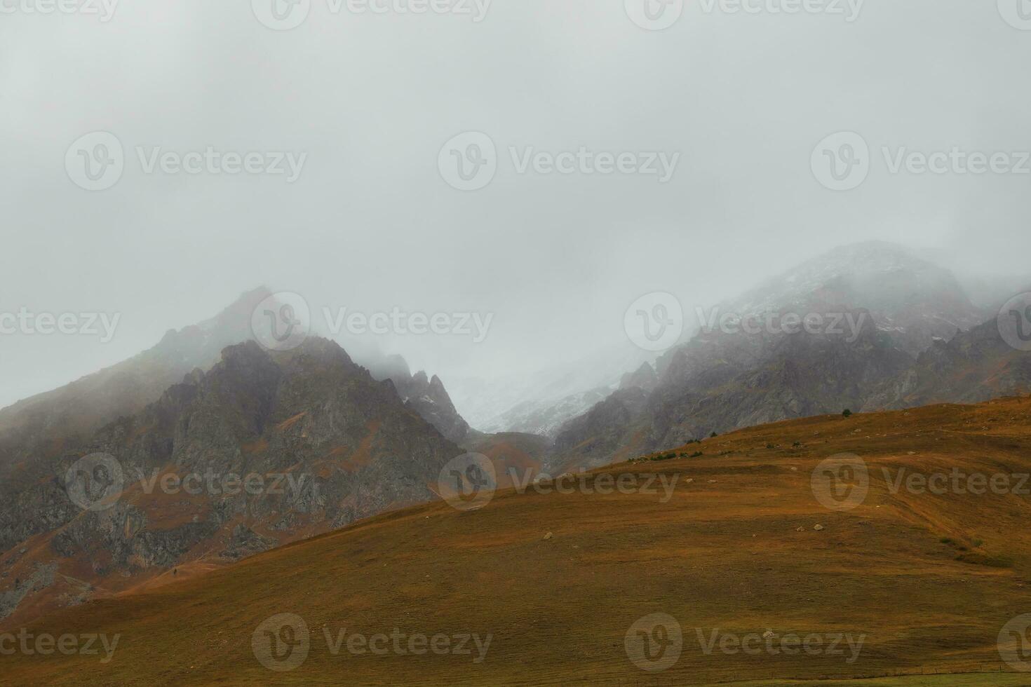 sfeervol nevelig landschap met wazig silhouetten van scherp rotsen in laag wolken gedurende regen. dramatisch visie naar groot bergen wazig in regen nevel in grijs laag wolken. foto