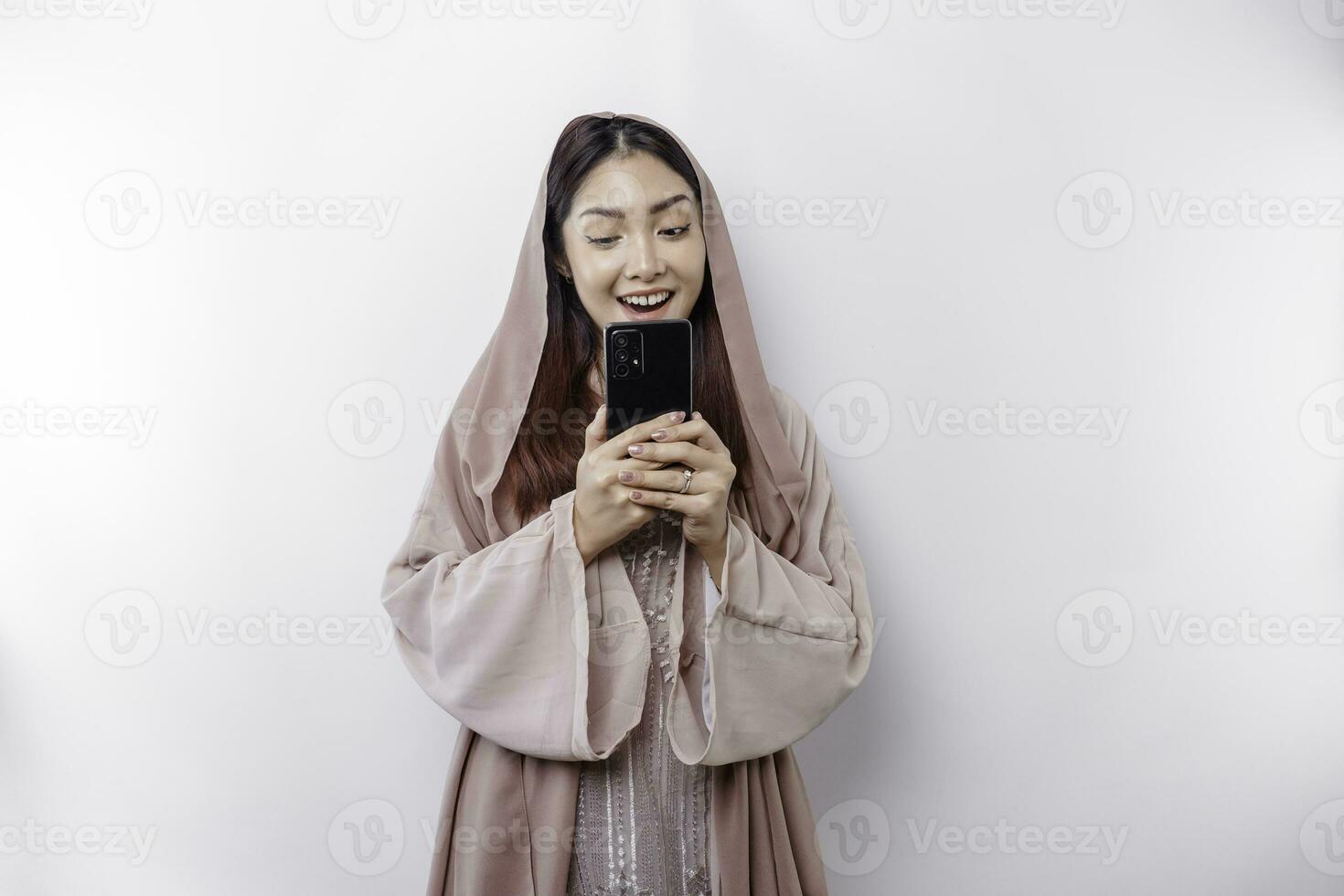 een gelukkig Aziatisch moslim vrouw vervelend een hoofddoek, Holding haar telefoon, geïsoleerd door wit achtergrond foto