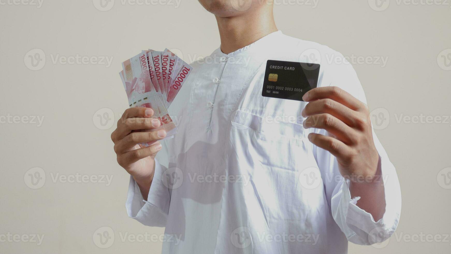 portret van aantrekkelijk Aziatisch moslim Mens in wit overhemd met kalotje Holding een honderd duizend roepia en presenteren credit kaart. geïsoleerd beeld Aan grijs achtergrond foto