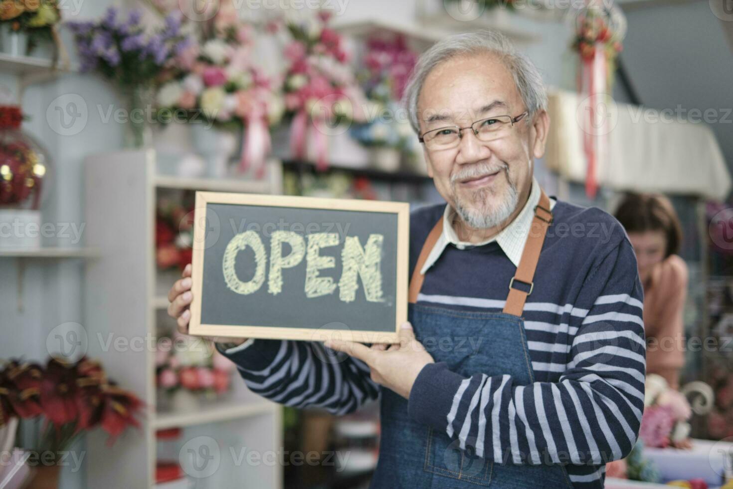 een senior mannetje bloemist eigenaar in schort shows Open teken bord in helder bloem winkel op te slaan met glimlach en looks Bij camera, klein bedrijf opening, bezetting pensioen, gelukkig ouderen mkb ondernemer. foto