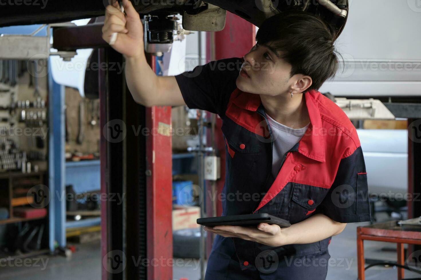 professioneel jong Aziatisch mannetje motor monteur inspecteert onderstel van elektrisch auto opgeheven door heftruck jack voor reparatie Bij garage, automotive onderhoud onderhoud werken industrie bezetting bedrijf. foto