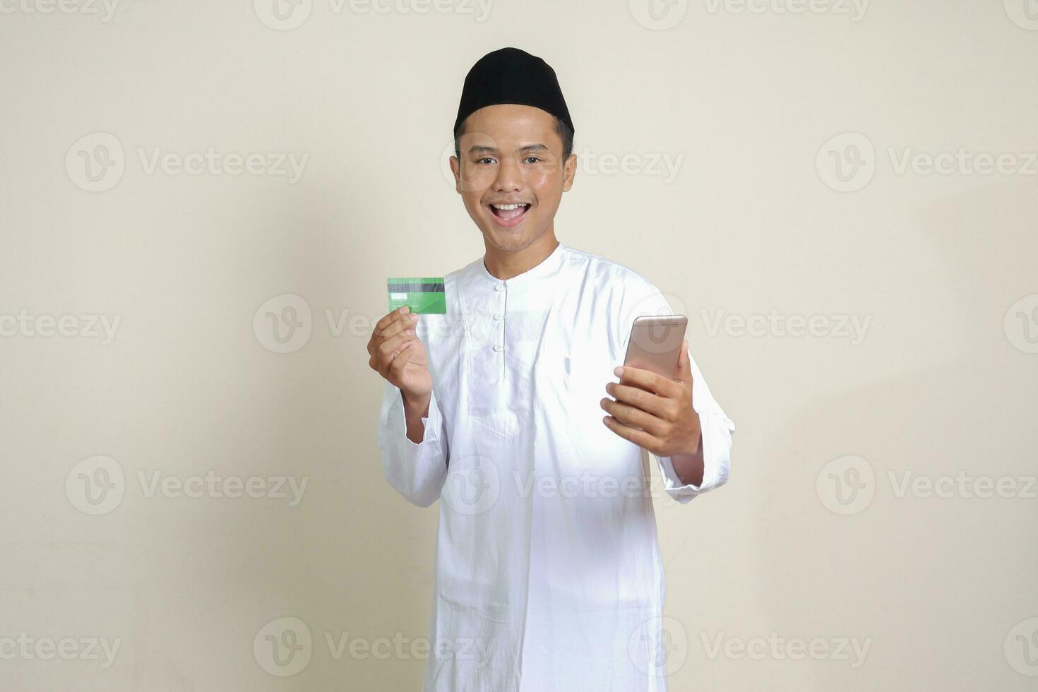 portret van aantrekkelijk Aziatisch moslim Mens in wit overhemd met kalotje Holding een mobiel telefoon en presenteren credit kaart. geïsoleerd beeld Aan grijs achtergrond foto
