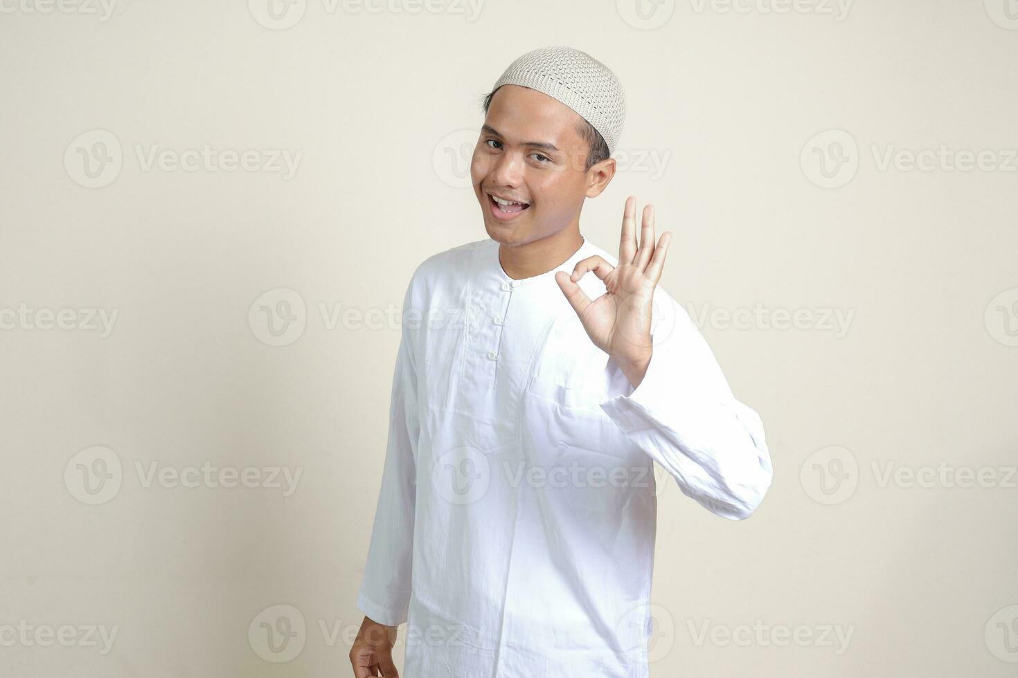 portret van aantrekkelijk Aziatisch moslim Mens in wit overhemd met kalotje tonen OK hand- gebaar en glimlachen op zoek Bij camera. reclame concept. geïsoleerd beeld Aan grijs achtergrond foto