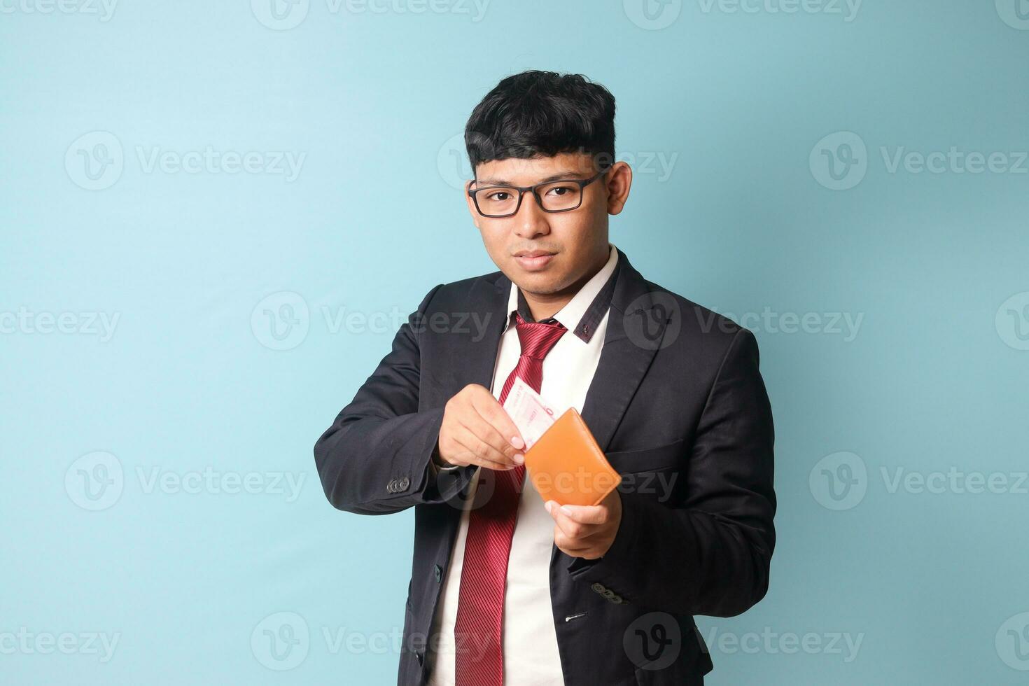 portret van jong Aziatisch bedrijf Mens in gewoontjes pak Holding leer portemonnee en trekken uit duizend roepia's terwijl op zoek Bij camera. geïsoleerd beeld Aan blauw achtergrond foto