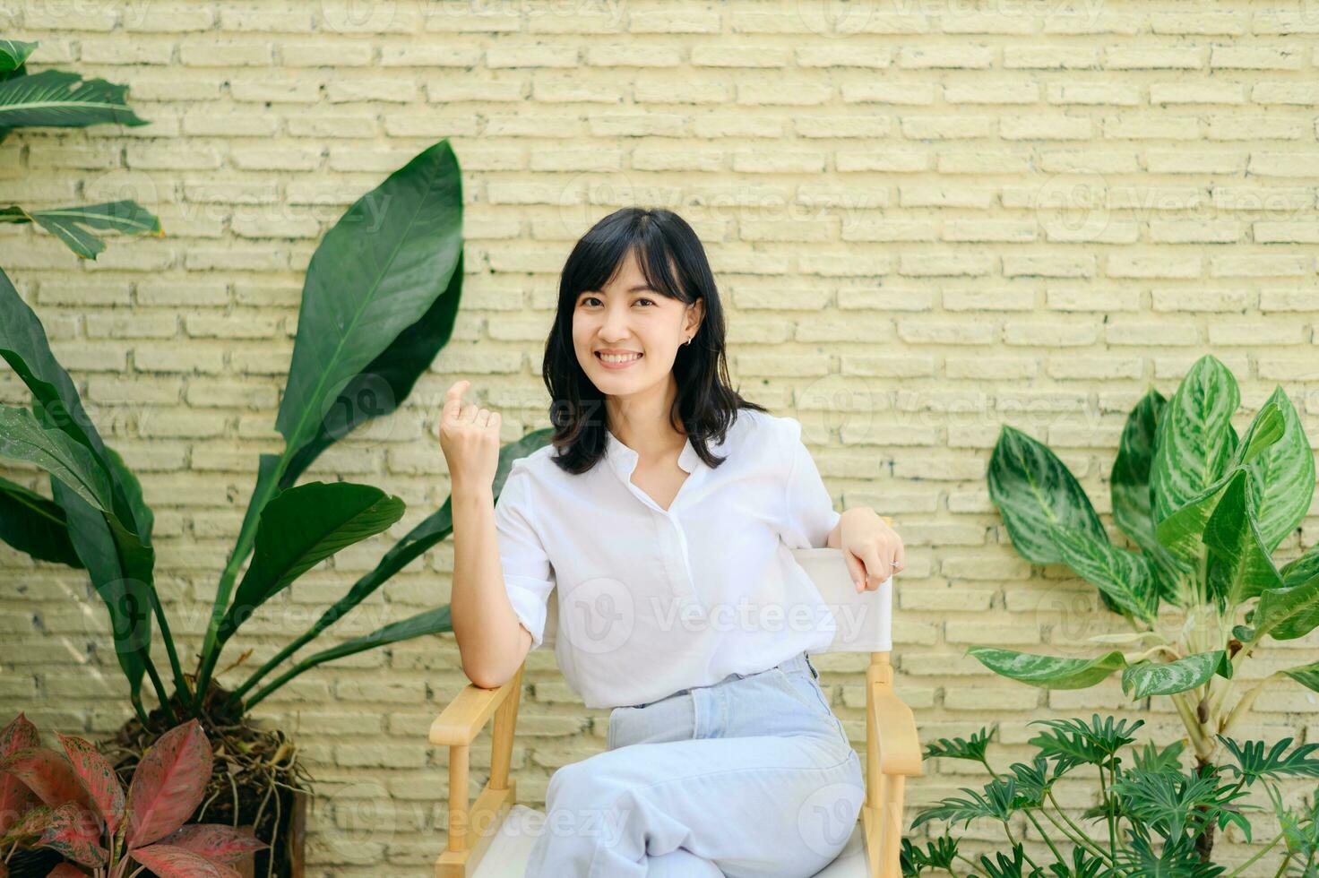 portret mooi jong Aziatisch vrouw geluk zittend Aan geel cement structuur grunge muur steen en groen fabriek achtergrond, gelukkig vrouw is glimlachen Aan concreet, welzijn mensen concept. foto