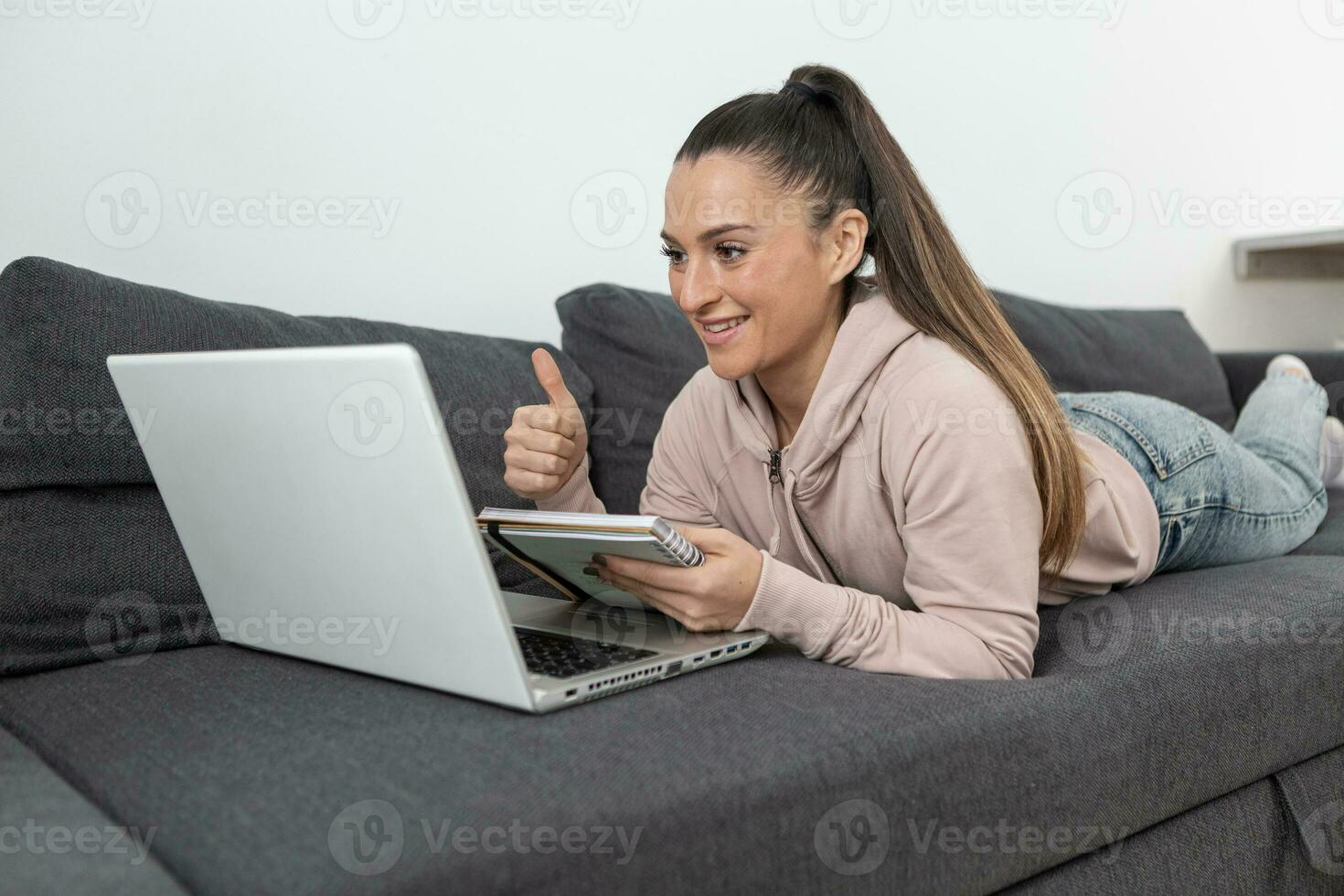 glimlachen vrouw aan het liegen Aan sofa met duimen omhoog nemen aantekeningen Aan laptop computer foto