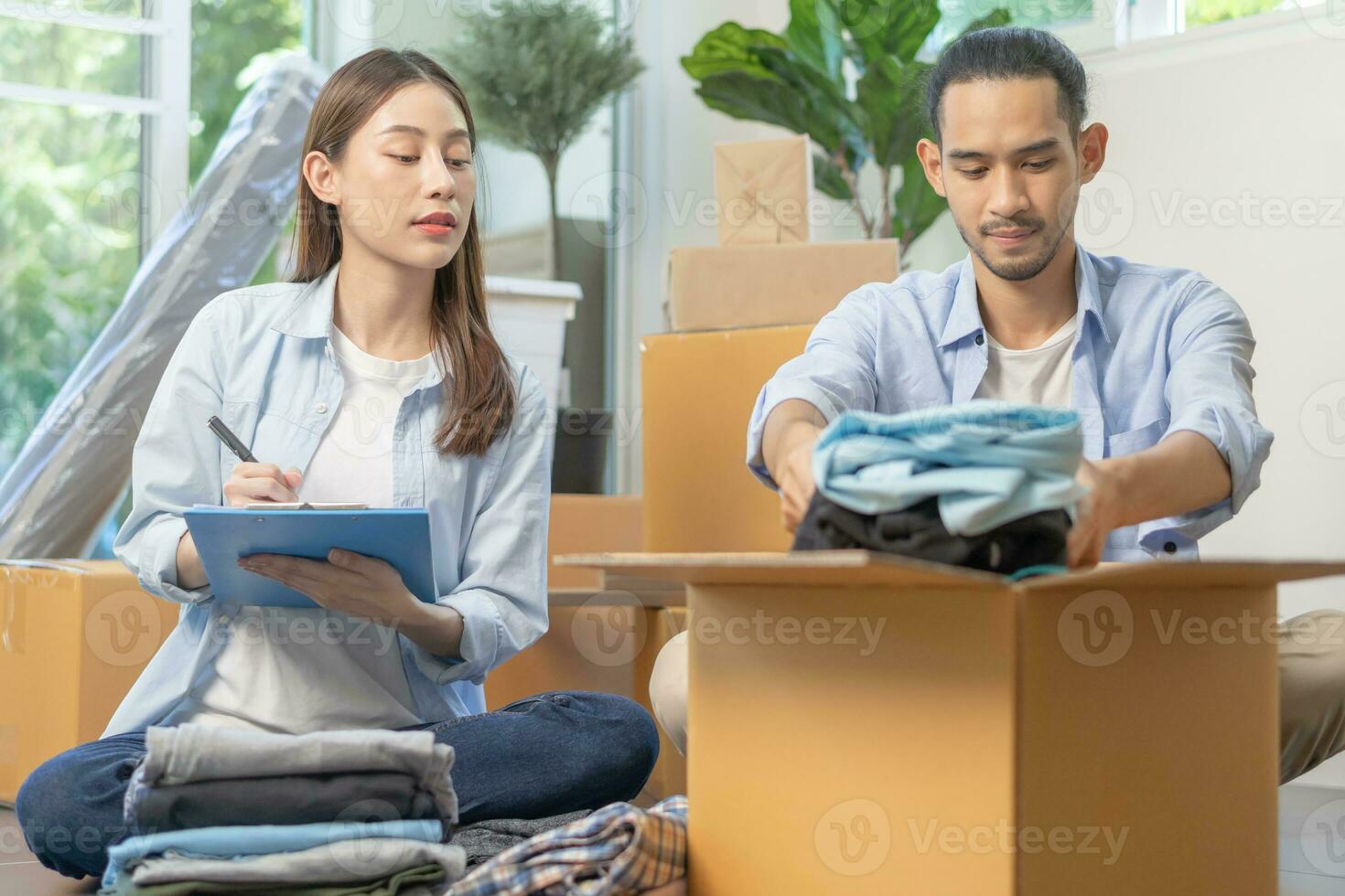 gelukkig paar Aziatisch jong vrouw, man inpakken dingen kleren, geven tweede hand- voor doneren, controleren lijst voorwerp in karton doos. in beweging, voorbereidingen treffen in nieuw huis, appartement verhuizing. foto