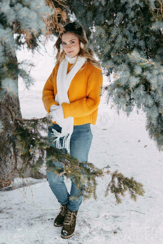 tiener blond in een geel trui buiten in winter. een tiener- meisje Aan een wandelen in winter kleren in een besneeuwd Woud foto