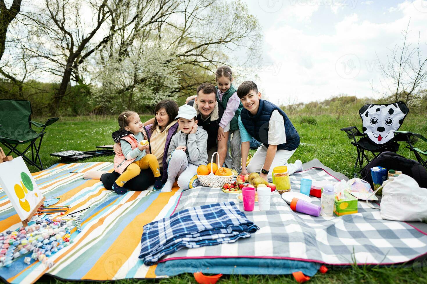 gelukkig jong groot familie met vier kinderen hebben pret en genieten van buitenshuis Aan picknick deken Bij tuin voorjaar park, ontspanning. foto