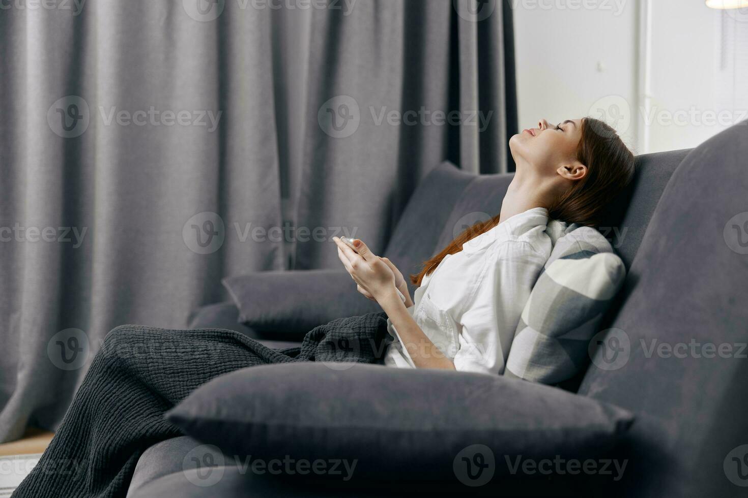 vrouw met mobiel telefoon Aan bankstel op zoek omhoog en gordijnen in de achtergrond foto