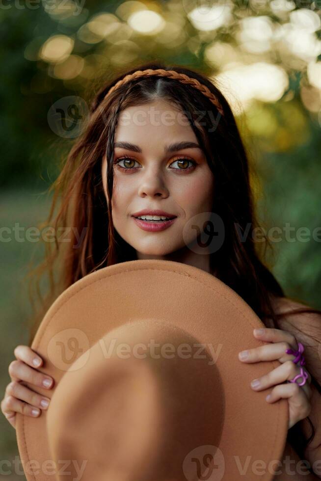 jong hippie vrouw Holding een hoed en glimlachen vriendelijke groet terwijl op zoek Bij de camera in natuur in de vallen foto
