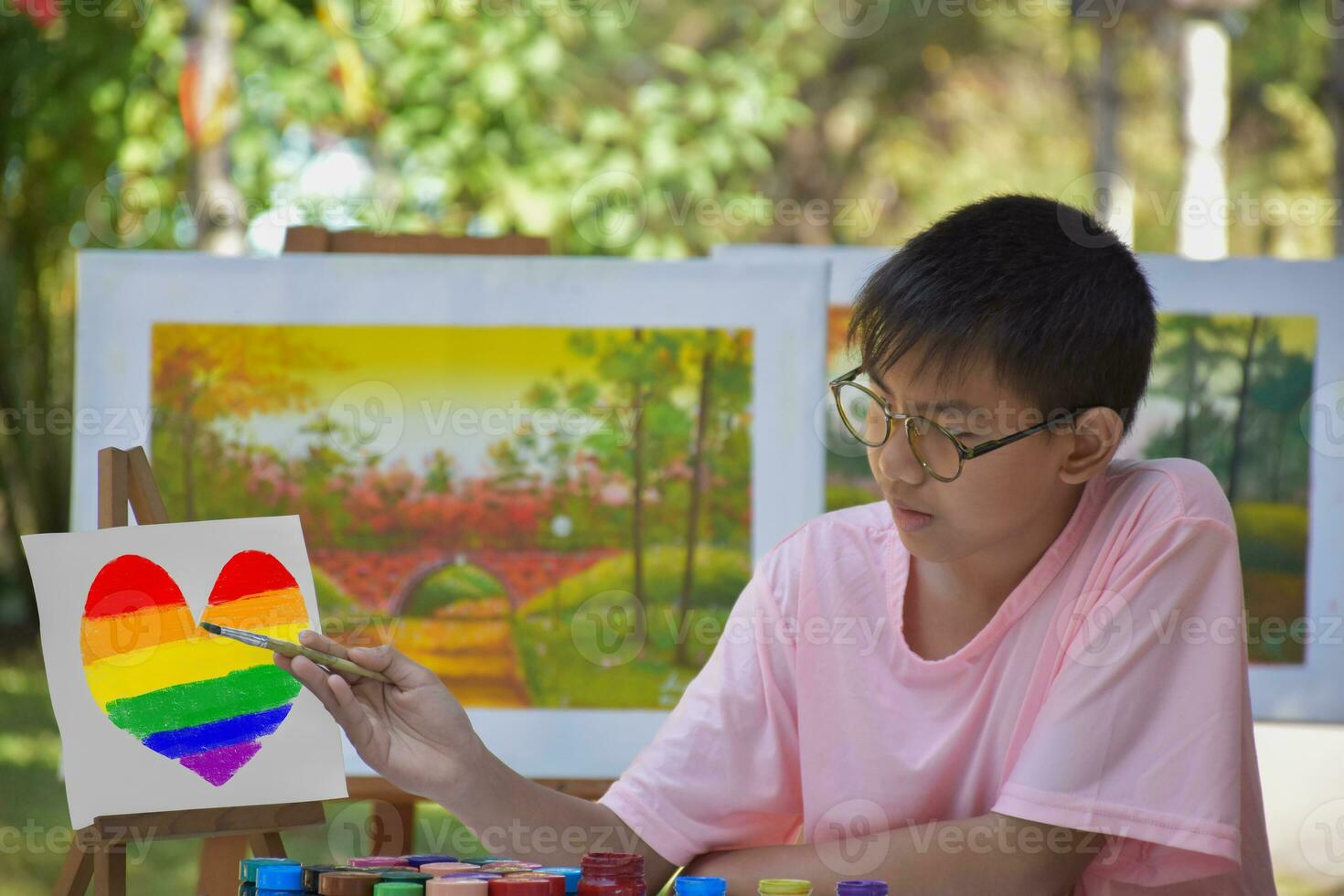 Aziatisch jongen in roze t-shirt uitgeven zijn vrije tijd gedurende zomer met buitenshuis kunst waterverf en gipskleur tekening, hij is tekening en kleur regenboog hart door gebruik makend van kleuren en borstel vooraan van hem. foto