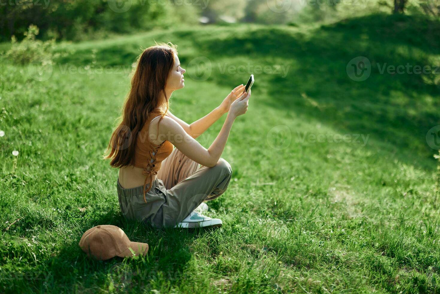 een jong gelukkig vrouw zittend in de park Aan de groen gras in natuur met haar telefoon in haar handen in haar gewoontjes kleren en chatten via video koppeling, de concept van gezond recreatie meditatie foto