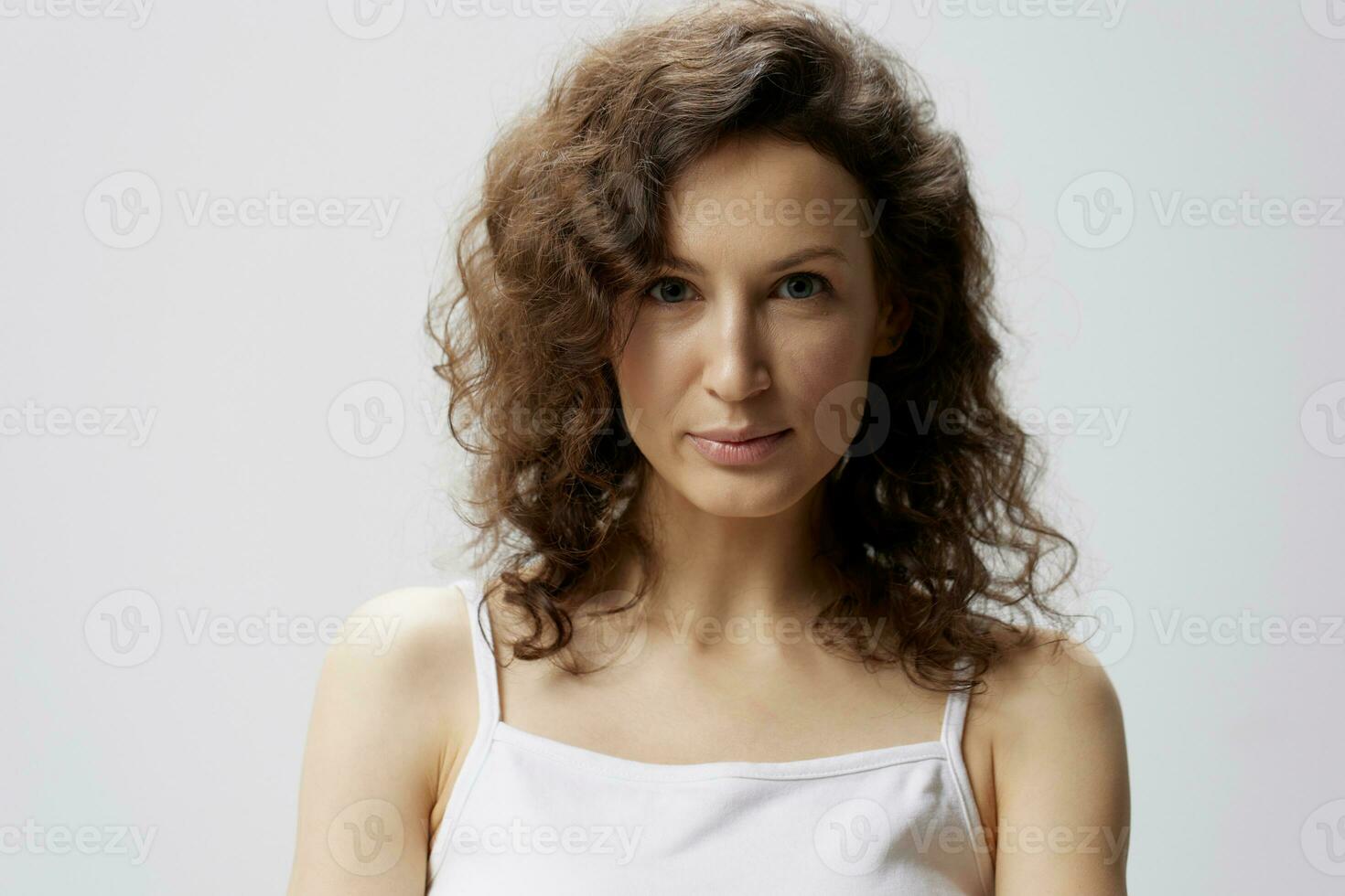 nadenkend lastig glimlachen gekruld mooi vrouw in eenvoudig wit t-shirt looks Bij camera poseren geïsoleerd Aan over- wit achtergrond. mensen emoties levensstijl concept. kopiëren ruimte. detailopname portret in studio foto