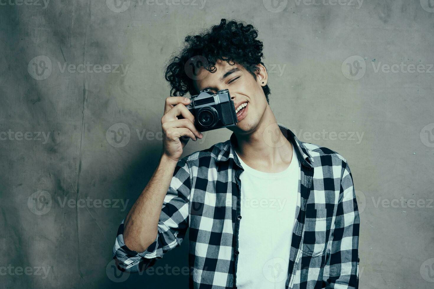 jong Mens met een camera in hand- en in een plaid t-shirt Aan een grijs achtergrond binnenshuis fotograaf foto