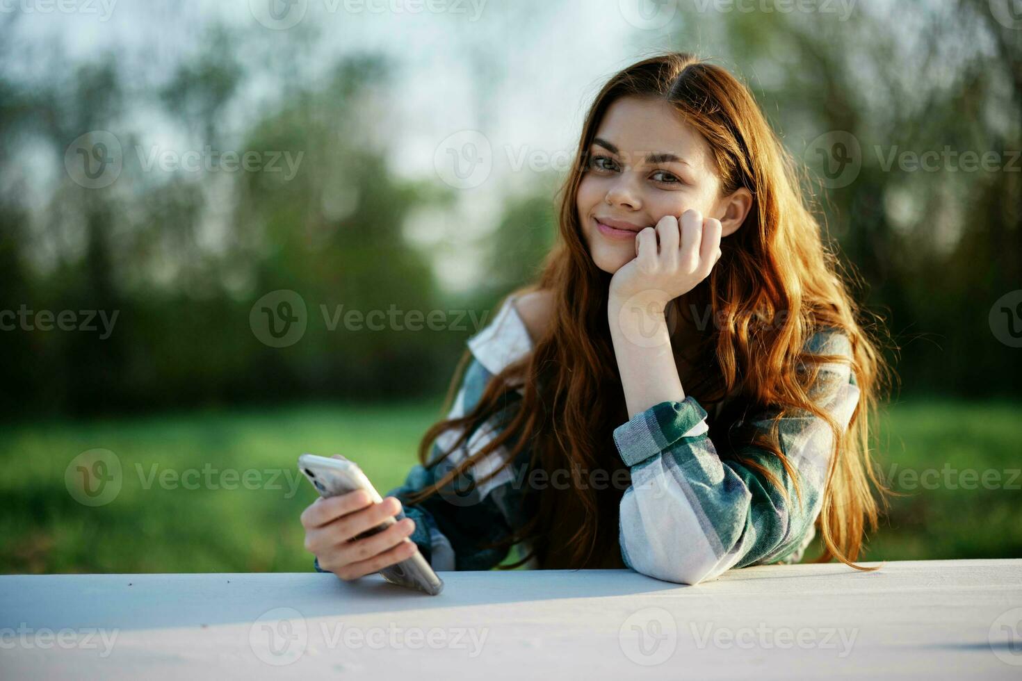 een vrouw met gekruld rood haar- in een plaid overhemd zit buitenshuis in de park met een cel telefoon in haar handen en looks weg. freelance werk in de park foto