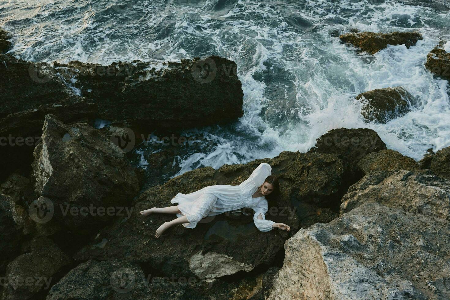 blootsvoets vrouw in lang wit jurk nat haar- aan het liegen Aan een rotsachtig klif ongewijzigd foto
