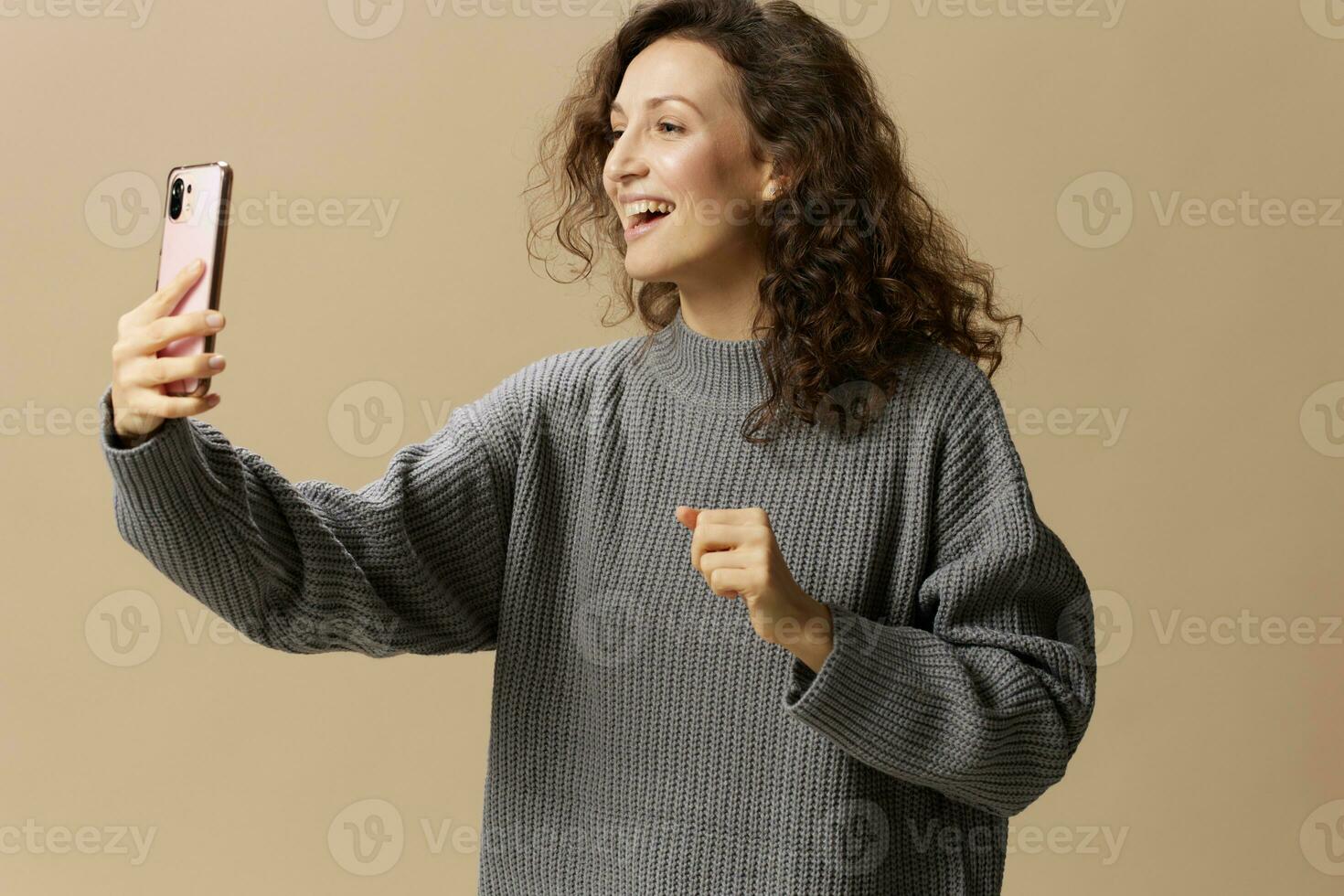 vrolijk opgewonden gekruld mooi vrouw in grijs gewoontjes trui Vermelding leven met telefoon poseren geïsoleerd Aan over- beige pastel achtergrond. sociaal media, netwerk, afstand communicatie concept. kopiëren ruimte foto