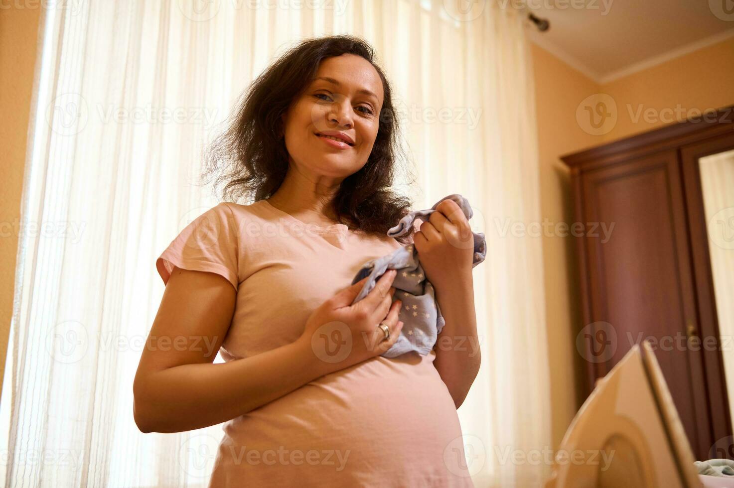 de verwachtend moeder is aan het wachten en voorbereidingen treffen voor de geboorte van een kind Bij huis, glimlachen Bij camera, Holding baby bodysuit foto