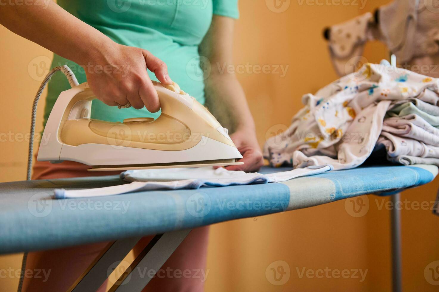 detailopname beige elektrisch stoom- ijzer in zwanger vrouw hand, strijken en stomen pasgeboren wasserij en baby kleren. foto