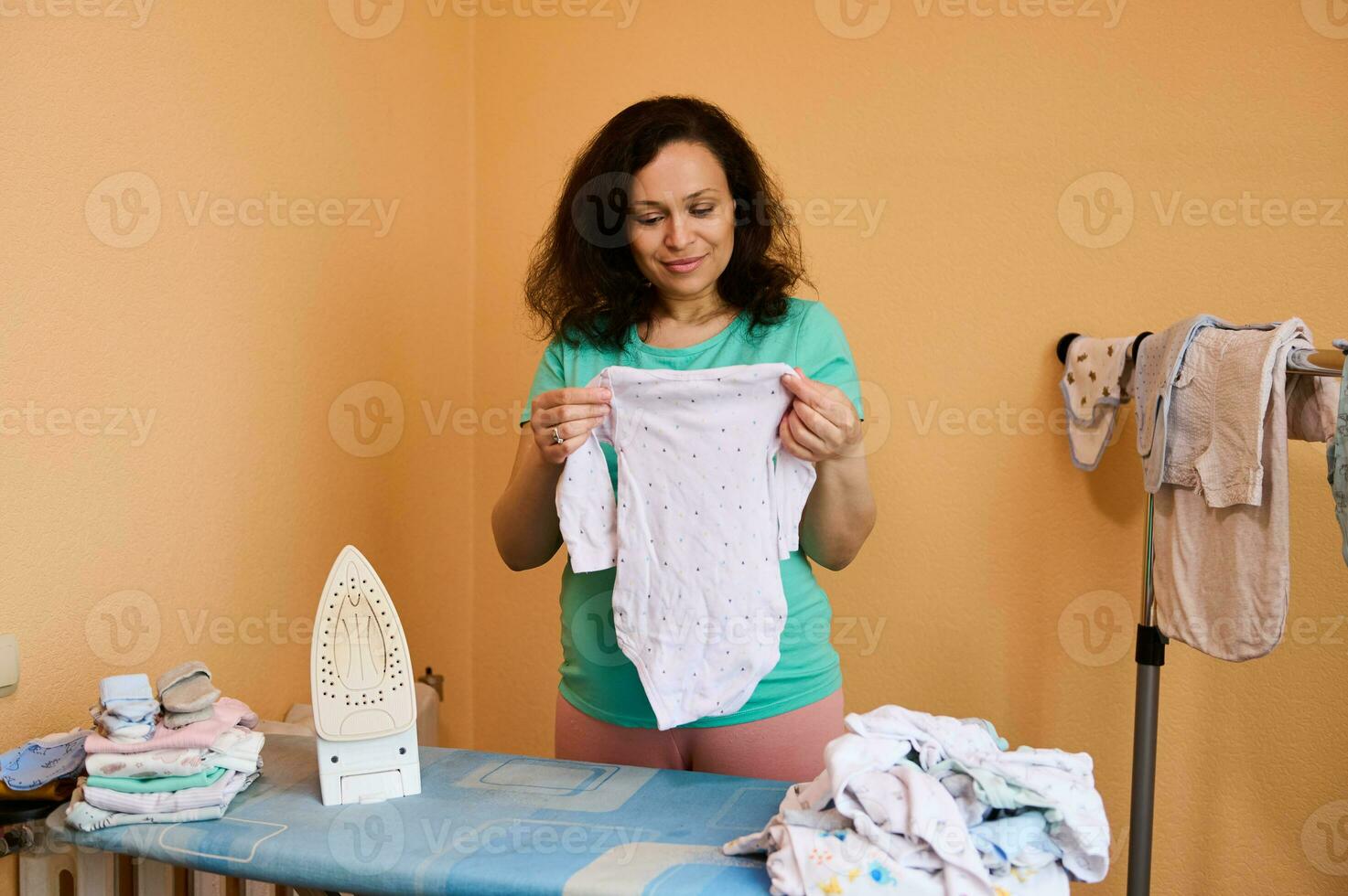 mooi zwanger vrouw, verwachtend mam Holding een witgewassen baby bodysuit, strijkijzers verfrommeld pasgeboren kleren Bij huis. foto