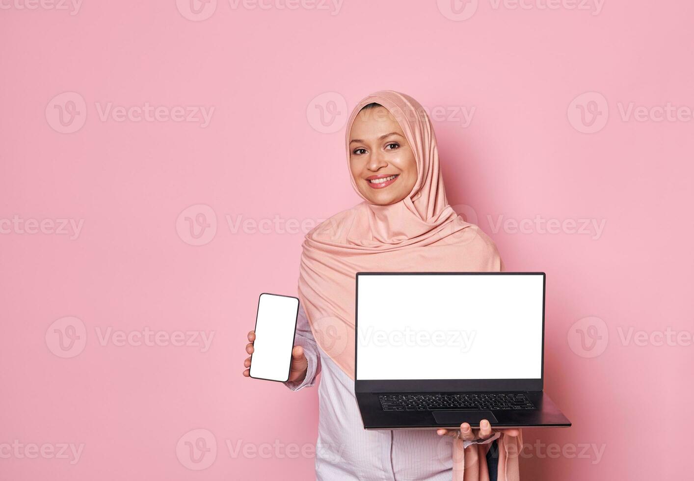 Arabisch moslim mooi vrouw in roze hijaab, glimlacht op zoek Bij camera, shows digitaal gadgets met wit blanco scherm. mockup foto