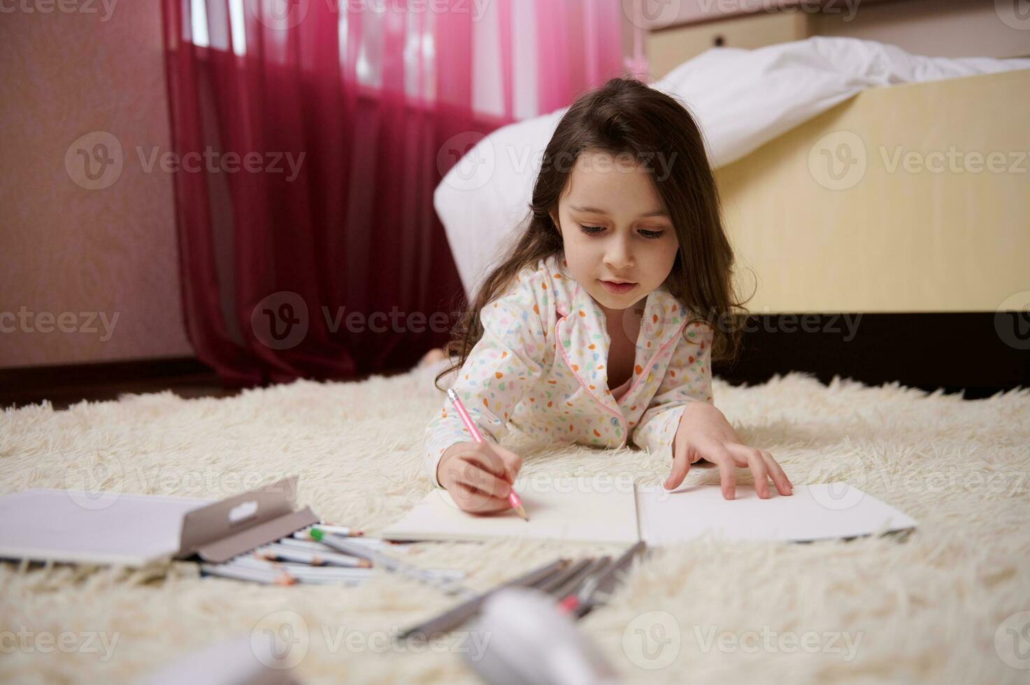 lief weinig meisje in pyjama, aan het liegen naar beneden Aan tapijt in slaapkamer, geconcentreerd Aan tekening afbeelding met kleurrijk potloden foto